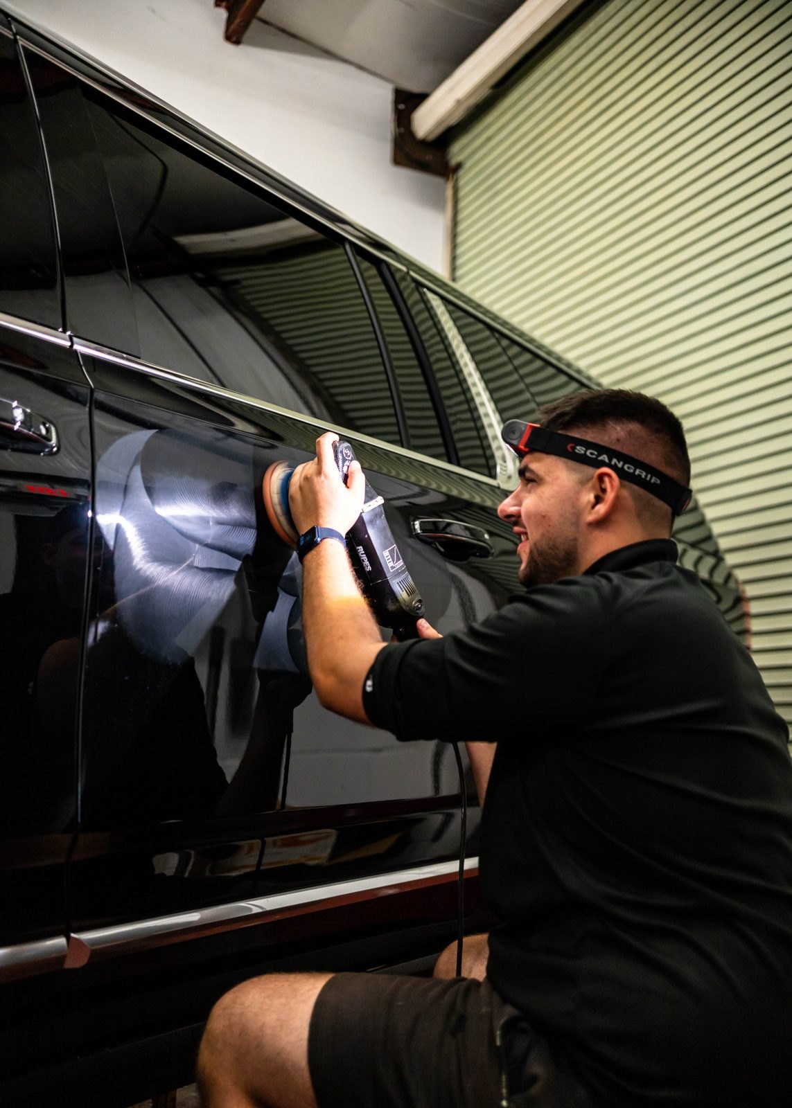 Detailer wearing a headlamp, running a polisher over the rear door of a black SUV in front of the shop's roll-up door