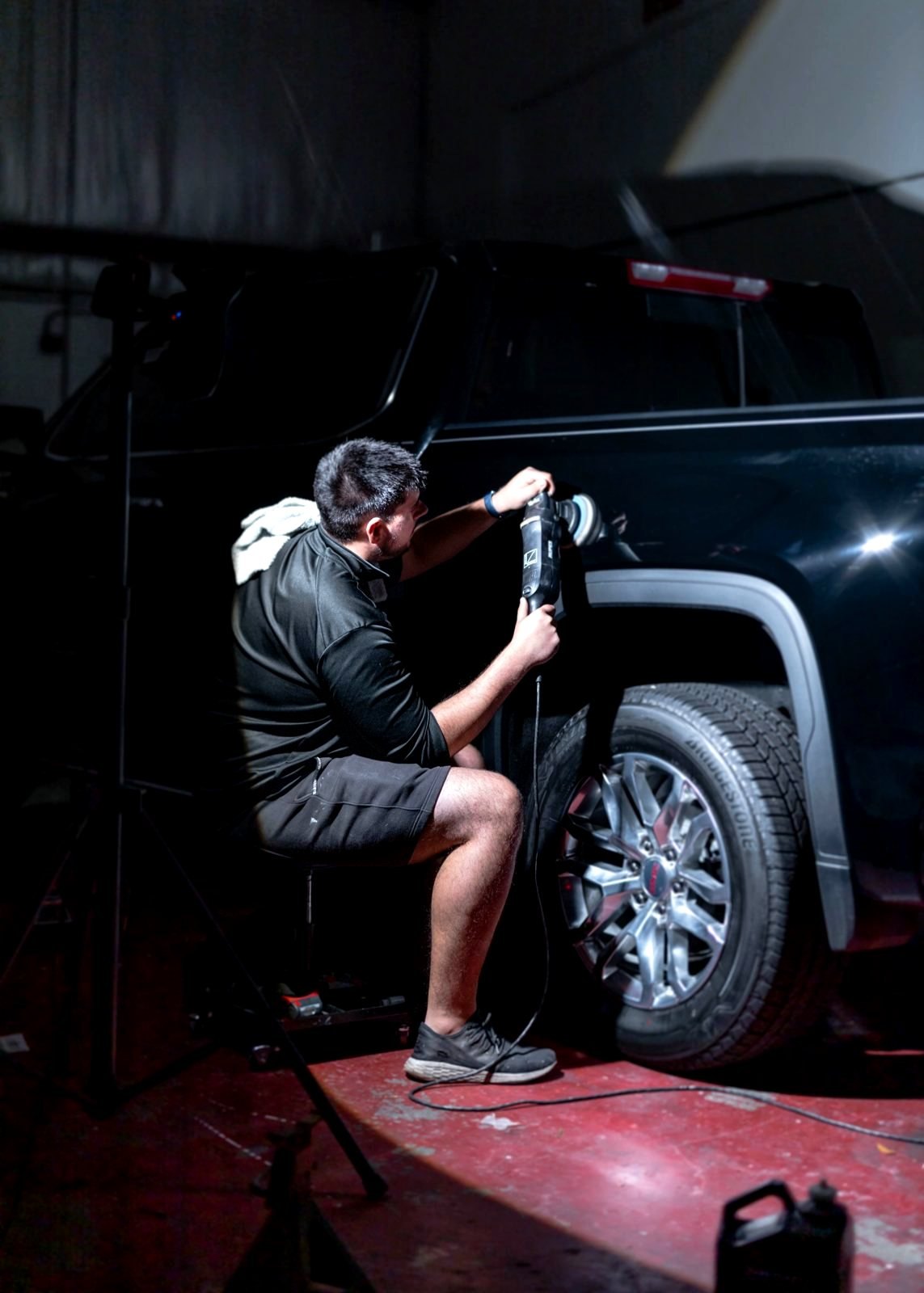 Detailer seated on a low stool, machine-polishing the side of a black pickup truck above the rear wheel in a dark bay
