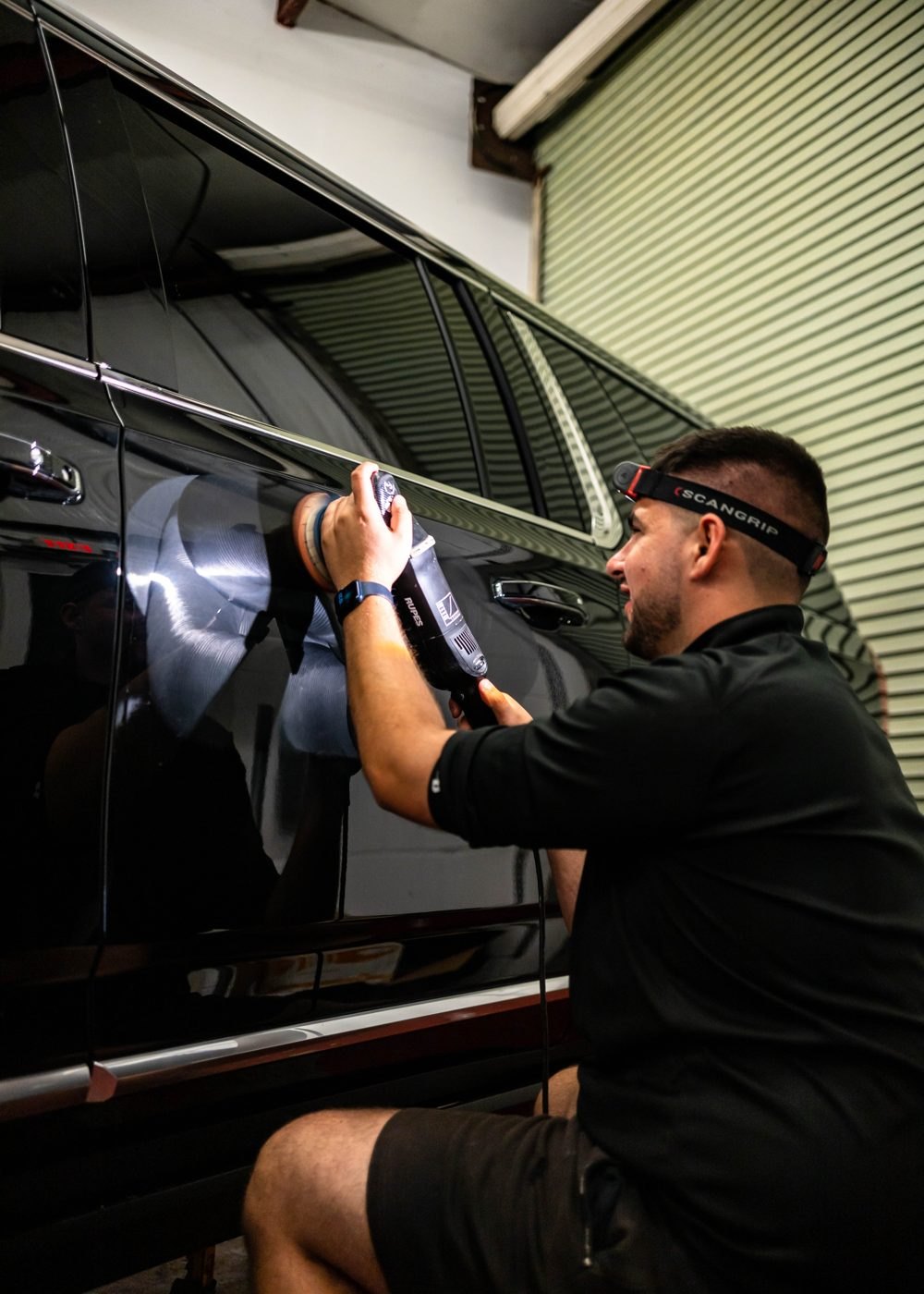 Detailer wearing a headlamp, machine-polishing the black door of an SUV beside its tinted window
