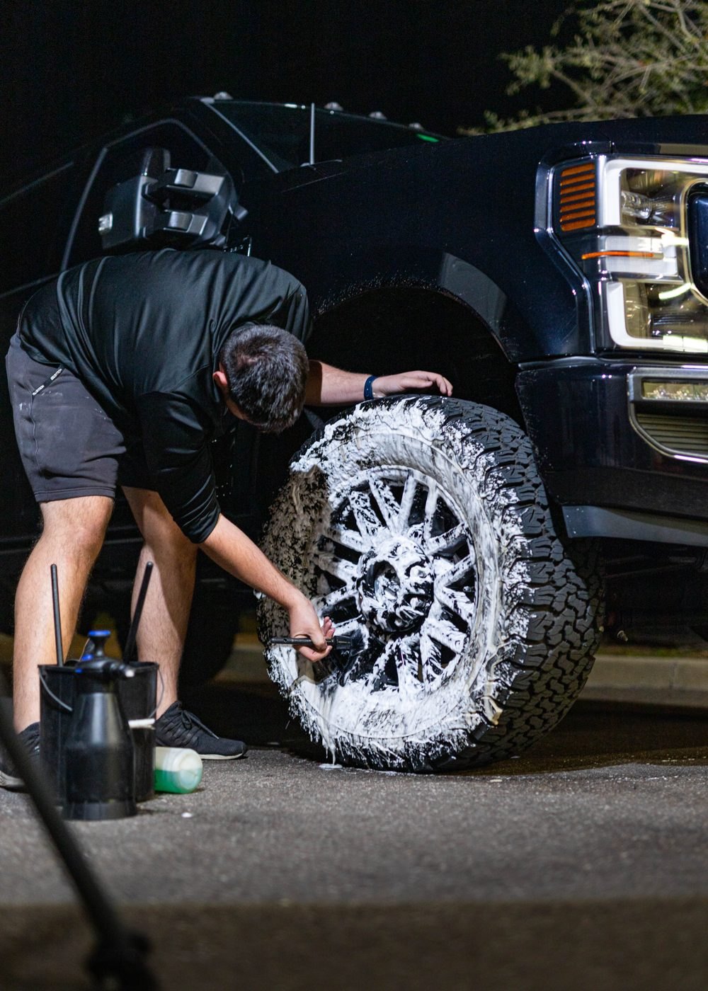 Detailer bent over the front wheel of a lifted black truck, scrubbing foam off the rim in a parking lot at night