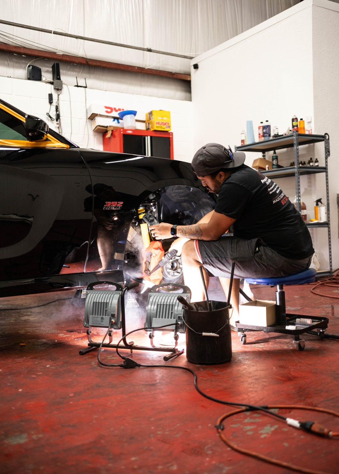 Detailer on a rolling stool working the front end of a black car under two work lights, with a shelf of bottles on the shop wall