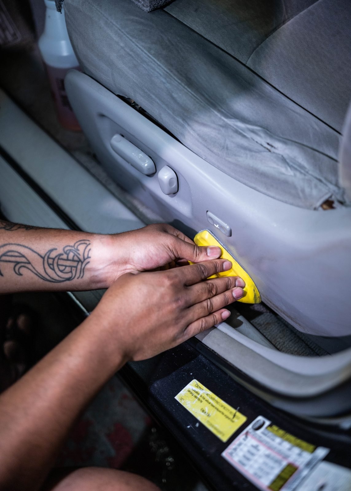 Hand working a yellow applicator pad along the plastic base of a car seat, with a bottle of cleaner on the floor behind it