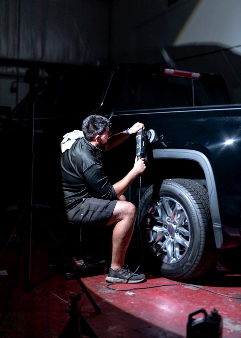 Detailer on a low stool machine-polishing the rear fender of a black pickup under a single work light